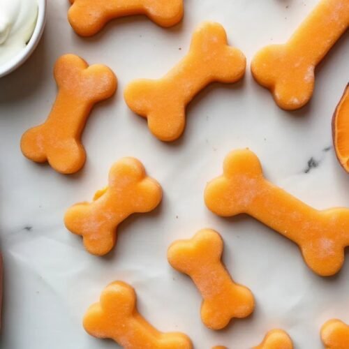 Overhead photo of homemade frozen dog treats shaped like bones, made with sweet potato and yogurt. The treats are pale orange and arranged on a light surface next to a bowl of Greek yogurt, a halved cooked sweet potato.
